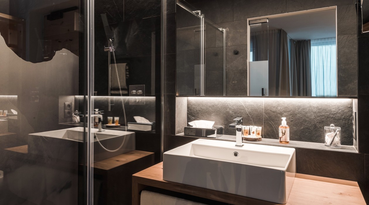 Modernes Badezimmer mit großer Regendusche, stilvollem Waschbecken, Natursteinwand und beleuchtetem Spiegel – Teil des Doppelzimmers im Hotel Panoramic Lotsch, Sarntal.