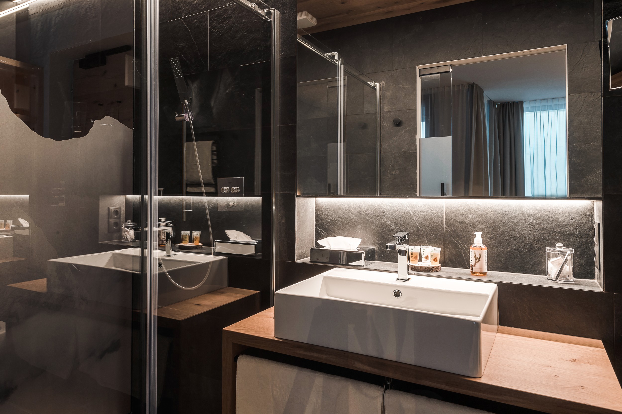 Modernes Badezimmer mit großer Regendusche, stilvollem Waschbecken, Natursteinwand und beleuchtetem Spiegel – Teil des Doppelzimmers im Hotel Panoramic Lotsch, Sarntal.