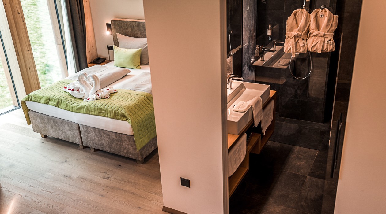 Hotel room with large window, double bed with green blanket and view into the modern bathroom at Hotel Panoramic Lotsch, Sarntal.