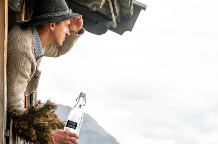 Südtiroler Tradition erleben – Mann mit Trachtenhut blickt aus urigem Berghüttenfenster mit Bergpanorama, Sarntal