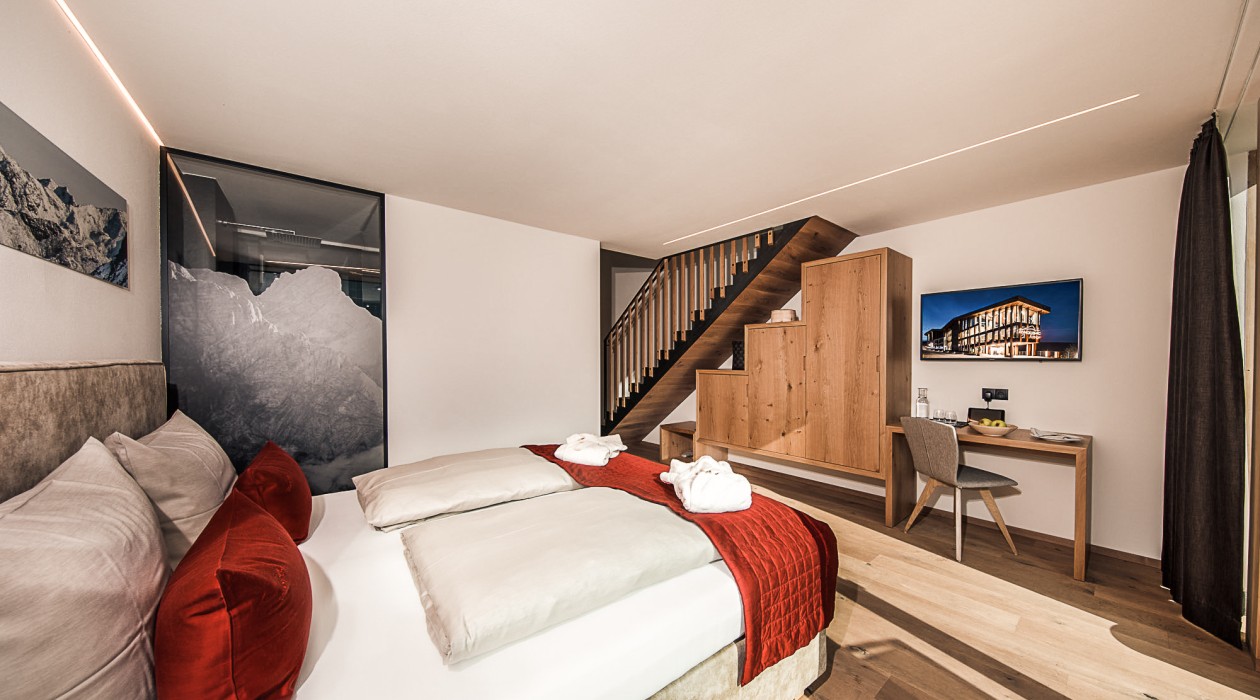 Schlafbereich und Wohnkomfort – Lotsch Suite im Hotel Panoramic Lotsch, Sarntal Gemütlicher Schlafbereich mit rotem Akzent, Schreibtisch und Holztreppe in der Lotsch Suite des Hotel Panoramic Lotsch, Sarntal – ruhige Wohnatmosphäre in Südtirol.