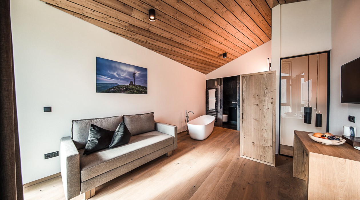 Sofa und Badewanne mit Design-Holzdecke – Lotsch Suite, Hotel Panoramic Lotsch, Sarntal Wohnbereich mit freistehender Badewanne, gemütlichem Sofa und Holzdecke in der Lotsch Suite des Hotel Panoramic Lotsch, Sarntal – moderner Rückzugsort in ruhiger Lage in Südtirol.