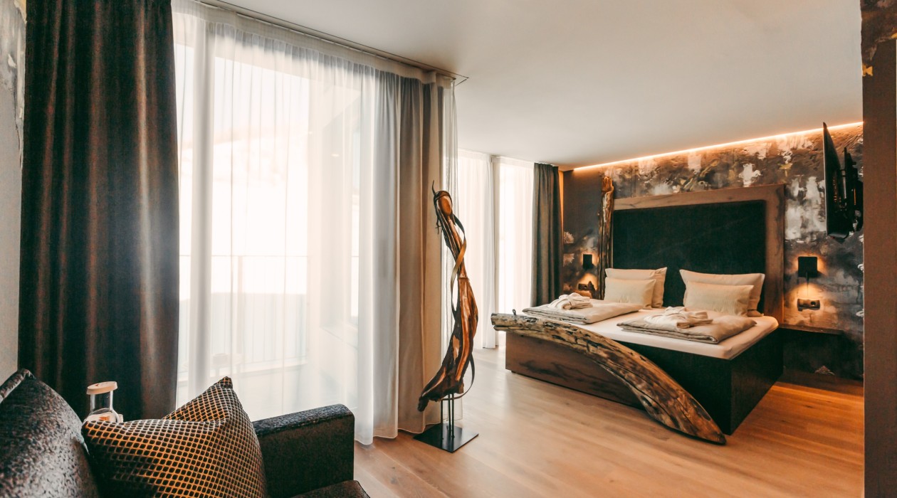 Double bed with carved wildwood element, artistic wall design and ambient lighting at Hotel Panoramic Lotsch, Sarntal – extraordinary room with peace and character in South Tyrol.