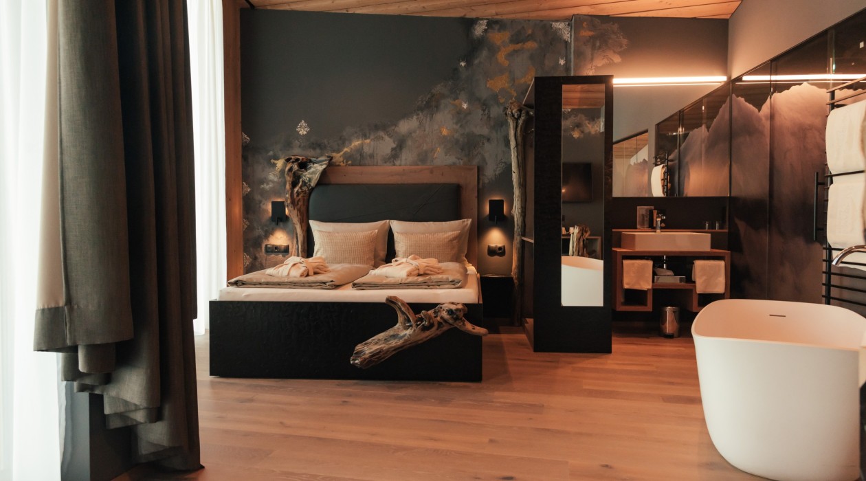 Suite mit Bett, Badewanne und Wildholz-Details – Hotel Panoramic Lotsch, Sarntal Wildholz-Suite mit Doppelbett, freistehender Badewanne, Naturholzelementen und künstlerischer Wandgestaltung im Hotel Panoramic Lotsch, Sarntal – stilvolle Ruhezone in Südtirol.
