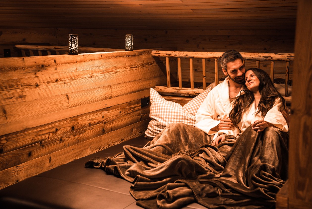 Paar entspannt in einer gemütlichen Wellness-Almhütte mit Decken und warmem Licht
