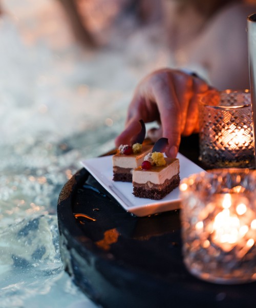 Hand greift nach einem Dessert auf einem Serviertablett am Rand eines Whirlpools mit Kerzenlicht